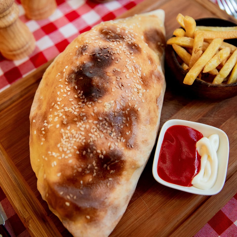 Golden-brown calzone with sesame seeds, fries, ketchup, and mayonnaise on a wooden board.