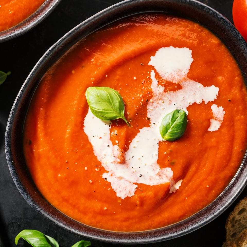 Two bowls of tomato soup with cream swirls, basil, fresh tomato, and bread slices.