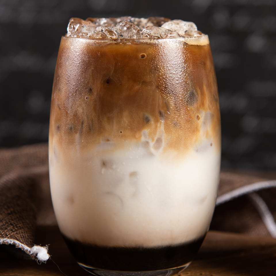 Iced coffee with milk and ice in a clear glass, with coffee beans on a wooden board.