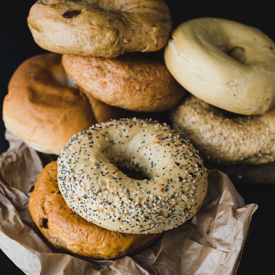 A pile of assorted bagels, including plain, sesame, poppy seed, and cinnamon raisin, on brown paper.