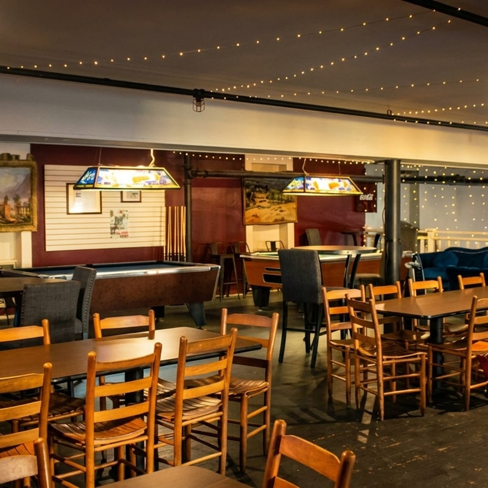 A dimly lit restaurant interior with string lights, wooden tables, chairs, and two pool tables.