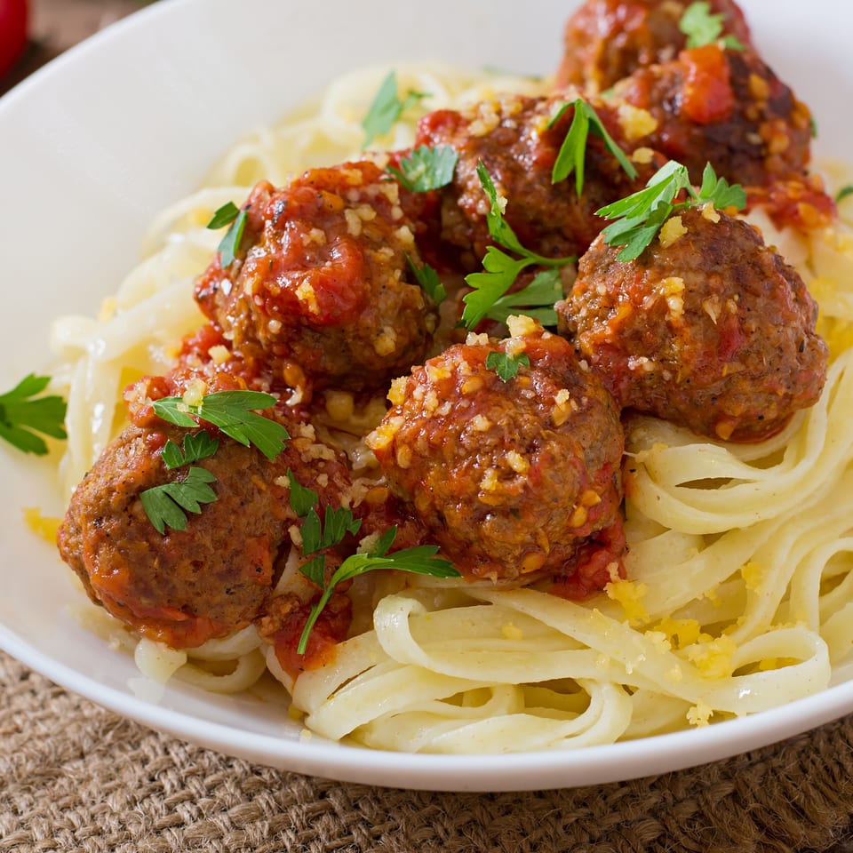 Close-up of spaghetti and meatballs in a white bowl, garnished with parsley and parmesan cheese.