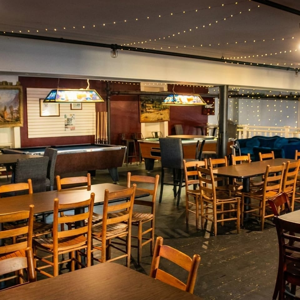 A dimly lit restaurant interior with wooden tables and chairs, pool tables, and string lights overhead.